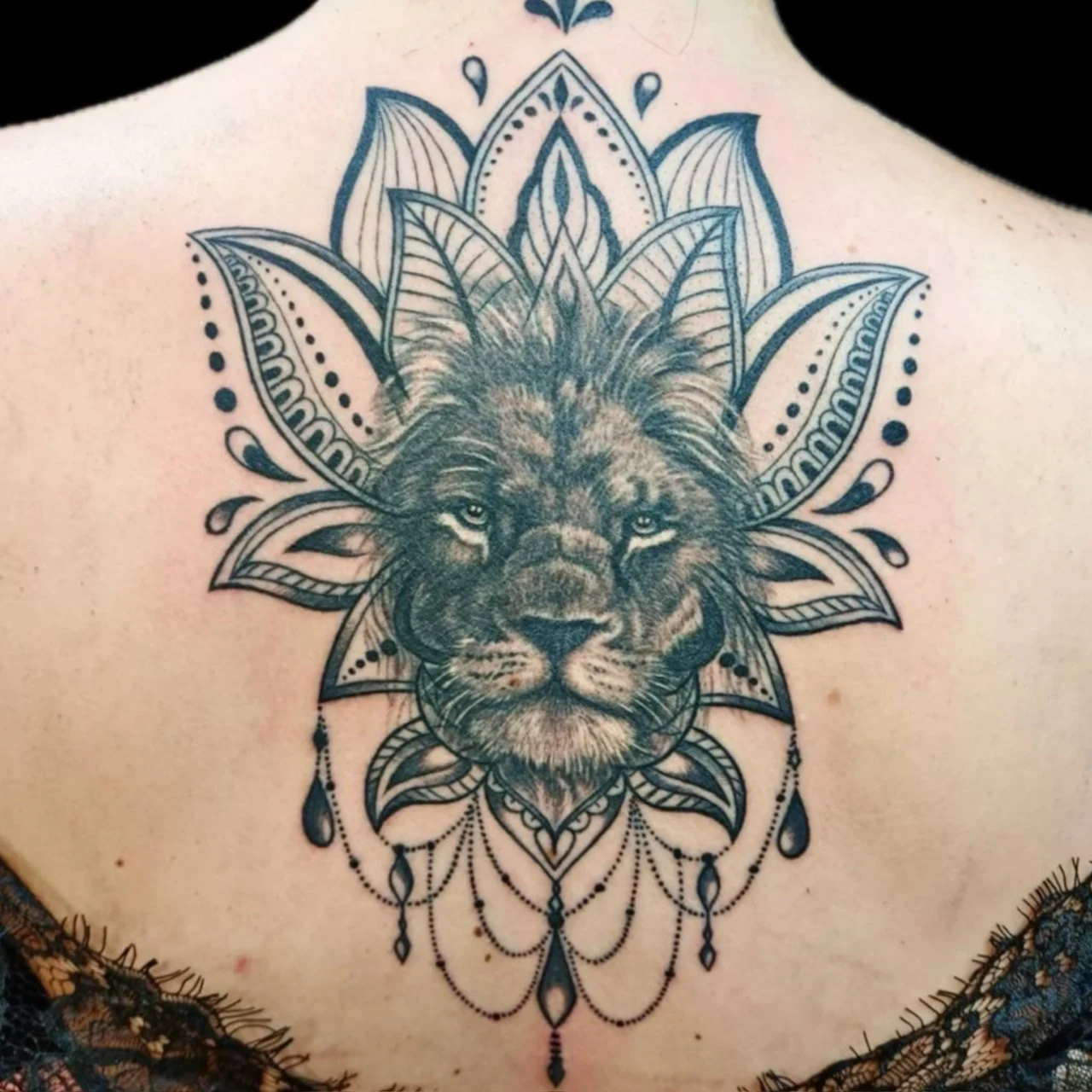 Ornamental upper-back tattoo with a realistic lion face centered in a patterned lotus mandala and hanging bead pendants at Bloodline Tattoo Bangkok.