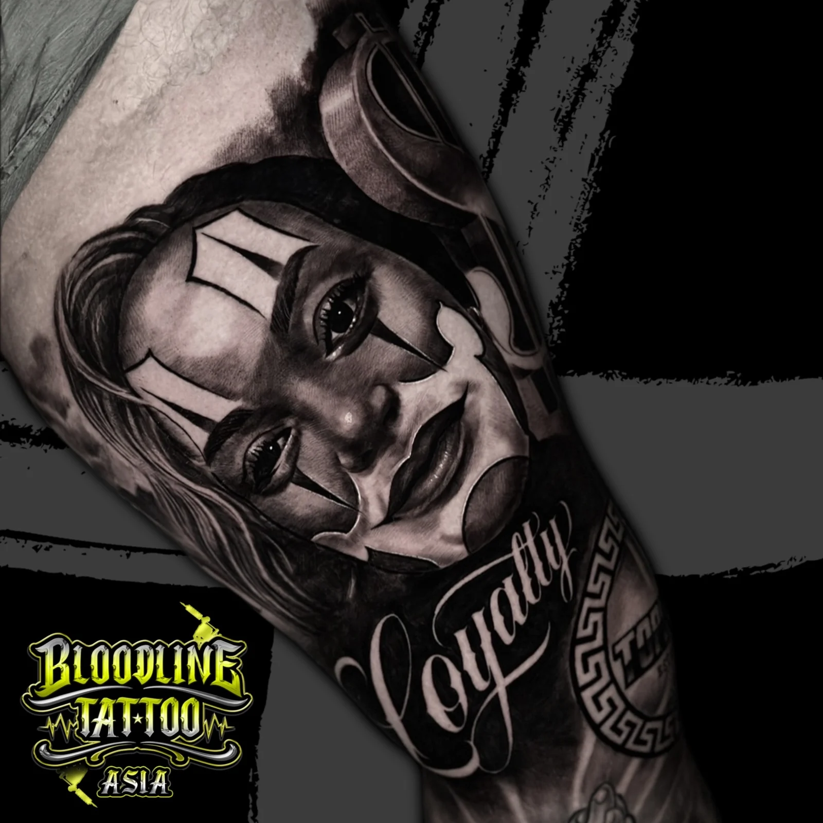 Black and grey Chicano arm tattoo with a payasa portrait, flowing Loyalty script, Greek key emblem, and praying hands at Bloodline Tattoo Bangkok.