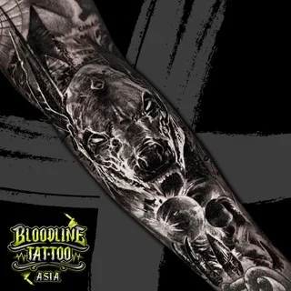 A black and grey realism sleeve tattoo of a snarling dog head with bared teeth and a "CANADA" landscape backdrop on the upper arm, at Bloodline Tattoo Bangkok.