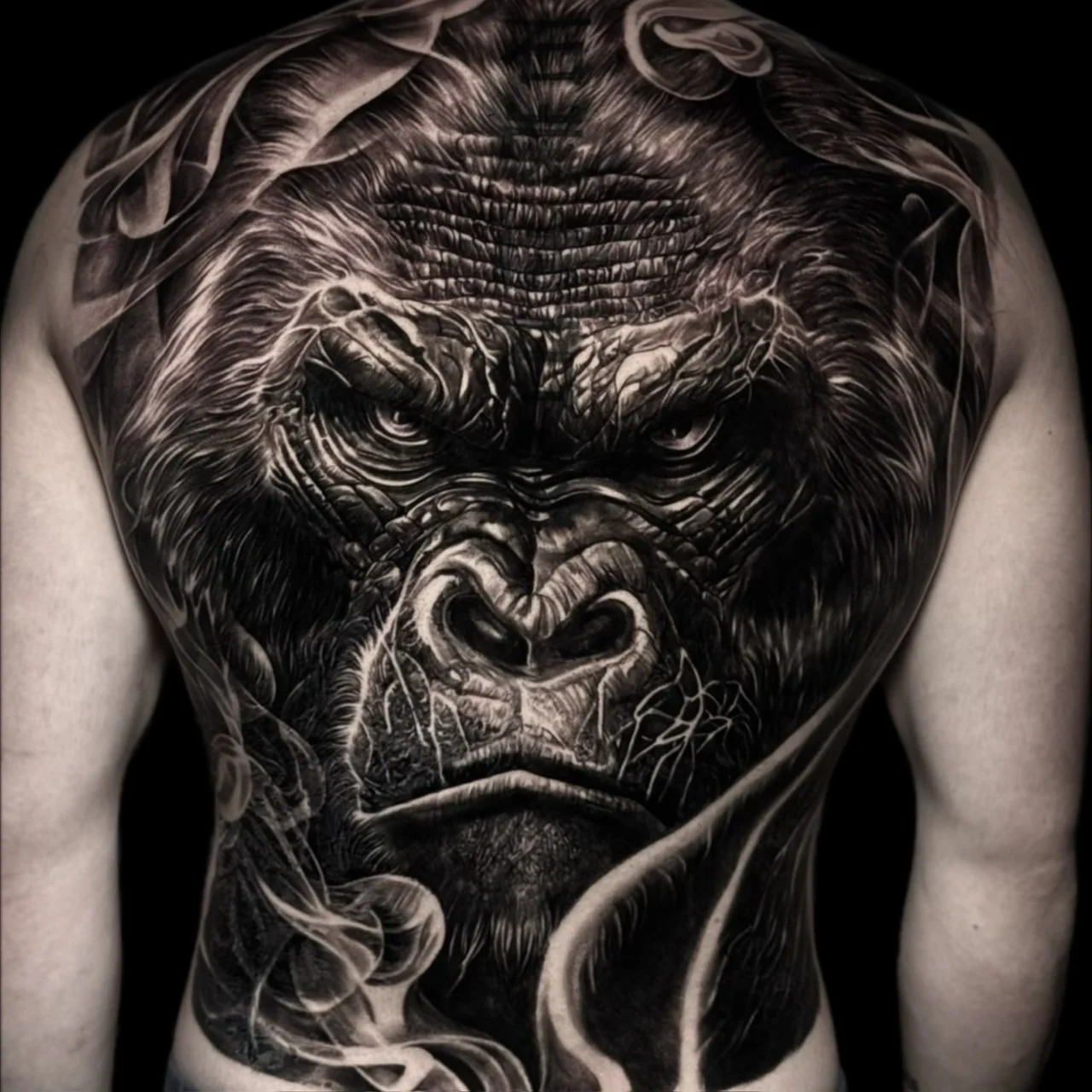 A black and grey realism full back piece of an intense gorilla portrait framed by smoky wisps across the shoulders and lower back, at Bloodline Tattoo Bangkok.